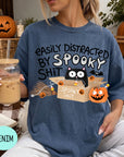 A woman wearing a blue t-shirt with a humorous Halloween-themed graphic, including a cat, pumpkins, and the text "EASILY DISTracted BY SPOOKY SHIT". She is holding a drink in a glass with a straw.