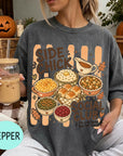 A woman wearing a gray t-shirt with a graphic design of various fall dishes and a pumpkin, holding a drink and surrounded by autumn decorations.