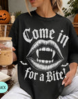 A woman wearing a black t-shirt with a graphic design of a pair of open-mouthed fangs and the text "Come in for a bite" is sitting on a couch with a pumpkin and a book nearby.