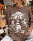 A woman is sitting on a couch, wearing a t-shirt with a Halloween-themed graphic design featuring characters such as Dracula, Frankenstein, and a ghost. She is holding a glass of iced coffee and has a book nearby.