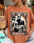 A woman wearing a t-shirt with a graphic design featuring a couple and the text "MONSTER THE LOVERS". She is holding a drink in a glass and there are Halloween decorations visible in the background.