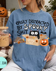 A woman wearing a blue t-shirt with a humorous Halloween-themed graphic, including a cat, pumpkin, and text that says "Easily Distracted by Spooky Shit". She is holding a drink in a glass with a straw.