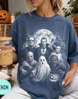 A woman wearing a blue t-shirt with a graphic design of a group of Halloween-themed characters, including Dracula, Frankenstein, Frankenstein's Bride, werewolf, and ghost, along with a pumpkin and other Halloween decorations. She is holding a drink in a glass with a straw.