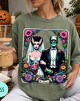 A woman wearing a green t-shirt with a graphic design of a bride and groom surrounded by flowers and a banner that says "The Lovers". She is holding a drink in a glass.