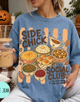 A woman wearing a blue t-shirt with a graphic design of various Thanksgiving dishes and the text "SIDE CHICK SOCIAL CLUB I'M JUST HERE FOR THE SIDES" is holding a drink and surrounded by fall decorations.