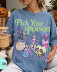 A woman wearing a blue t-shirt with a graphic design featuring various Halloween-themed items, such as a skull, a vase, and a bottle, along with the text "Pick Your Poison". She is holding a drink in a glass with a straw.