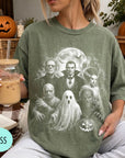 A person is sitting on a couch, wearing a green t-shirt with a Halloween-themed graphic of characters from the classic horror film "The Exorcist". The person is holding a drink in a glass with a straw.