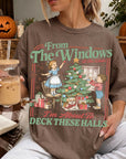 A woman wearing a t-shirt with a graphic design of a Christmas tree and two children decorating it, holding a drink and sitting on a couch with a book and a pumpkin decoration in the background.
