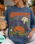 A woman wearing a blue t-shirt with a Halloween-themed graphic, holding a drink and surrounded by pumpkins and other fall decorations.