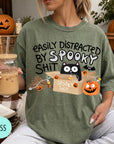 A person wearing a green t-shirt with a humorous Halloween-themed graphic and text, holding a drink and surrounded by Halloween decorations.