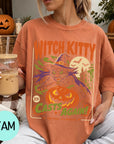 A woman wearing a t-shirt with a Halloween-themed design, including a witch, pumpkin, and bats, is holding a drink in her hand.