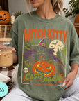 A woman wearing a green t-shirt with a Halloween-themed graphic and holding a drink, with a pumpkin and other fall decorations visible in the background.