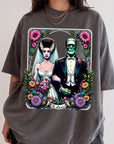 A graphic t-shirt featuring a bride and groom with a greenish-blue monster figure, surrounded by flowers and the text "The Lovers".