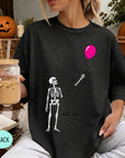 A woman wearing a black t-shirt with a skeleton and a pink balloon graphic, holding a drink and sitting on a blanket with a pumpkin and autumn leaves in the background.