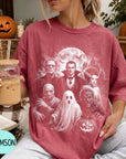 A woman wearing a red t-shirt with a Halloween-themed graphic of Dracula, Frankenstein, Frankenstein's Bride, Frankenstein's monster, and a ghost, holding a drink and sitting on a blanket.