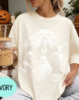 A woman wearing a white t-shirt with a Halloween-themed graphic, holding a glass of iced coffee, and surrounded by Halloween decorations.