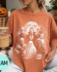A woman wearing a t-shirt with a Halloween-themed graphic of characters from the horror film "The Bride of Frankenstein" is holding a drink and sitting on a couch with a pumpkin and other Halloween decorations in the background.