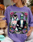 A woman wearing a purple t-shirt with a graphic design featuring a bride and groom, surrounded by flowers and a banner that says "The Lovers". She is holding a drink in a glass.