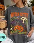 A woman wearing a gray t-shirt with a Halloween-themed graphic design, including a witch, pumpkin, and bats. She is holding a drink in a glass with a straw.