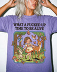 A woman wearing a purple t-shirt with a cartoon illustration of a young girl interacting with animals and nature, along with the text "WHAT A FUCKED UP TIME TO BE ALIVE".