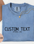 a blue denim t-shirt with the text "CUSTOM TEXT" printed on it, along with the phrase "OPTIONAL LINE" below it. The t-shirt is laid out on a white surface, accompanied by a beige hat, a gold chain, and some pink flowers.