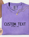 a purple t-shirt with the text "CUSTOM TEXT" and "OPTIONAL LINE" printed on it, along with a gold star charm and a beige hat. The t-shirt is laid out on a white surface, and there is a small pink circle with the word "VIOLET" written on it.