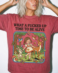 A woman wearing a red t-shirt with a cartoon illustration of a young girl interacting with animals, including a deer, rabbit, and birds, along with the text "WHAT A FUCKED UP TIME TO BE ALIVE".