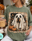 A woman is sitting on a couch, wearing a green t-shirt with a graphic of a ghost dog and pumpkins. She is holding a glass of iced coffee.