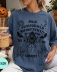 A woman wearing a blue t-shirt with a Ouija board design and holding a drink is sitting in front of a Halloween decoration.