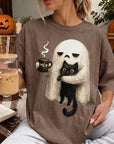 A woman is sitting on a couch, wearing a t-shirt with a graphic of a ghost holding a coffee mug and a black cat. She is holding a glass of iced coffee and a book.