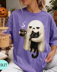 A person wearing a purple t-shirt with a graphic of a ghost holding a coffee mug and a black cat, sitting on a couch with a pumpkin and a book nearby.