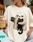 A person wearing a white t-shirt with a graphic of a ghost holding a coffee mug and a black cat, with a pumpkin and a book visible in the background.