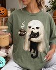 A person is sitting on a couch, wearing a green t-shirt with a graphic of a ghost holding a coffee mug and a cat.