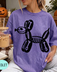 A woman wearing a purple t-shirt with a skeleton dog graphic is sitting on a blanket, holding a drink in a glass.