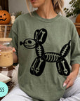 A woman wearing a green t-shirt with a skeleton dog graphic is sitting on a couch, holding a drink in a glass.