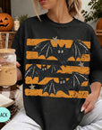 A woman wearing a black t-shirt with a graphic of bats and stars is sitting in a room with Halloween decorations, including a jack-o-lantern and a pumpkin. She is holding a drink in a glass with a straw.