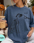 A woman wearing a blue t-shirt with a skeleton drawing and the word "boop" printed on it, holding a glass of iced coffee.