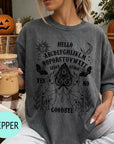 A woman wearing a gray t-shirt with a graphic design of an Ouija board and text printed on it, holding a drink in a glass.