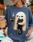 A woman wearing a blue t-shirt with a graphic of a ghost holding a coffee mug and a black cat, sitting on a couch with a pumpkin and a book nearby.