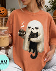 A woman wearing a t-shirt with a graphic of a ghost holding a coffee mug and a black cat, sitting on a couch with a pumpkin and a book nearby.
