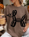 A woman is sitting on a blanket, wearing a t-shirt with a skeleton dog design, and holding a glass of iced coffee.