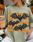 A woman is sitting on a couch, wearing a green t-shirt with a graphic of bats and stars. She is holding a glass of iced coffee.