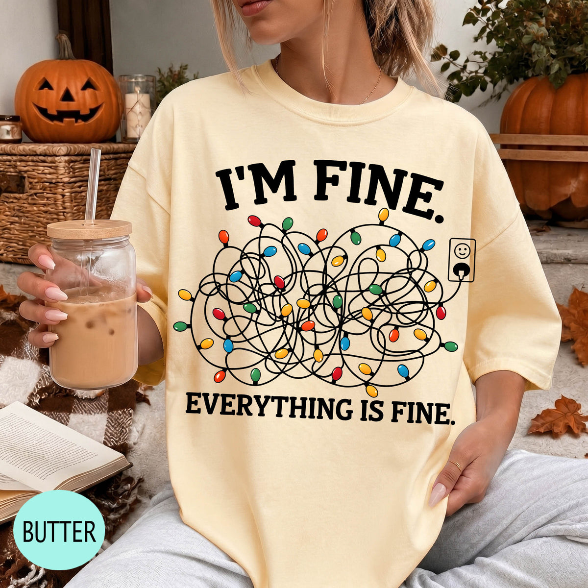 A person wearing a yellow t-shirt with a graphic design and text, sitting on a couch with a pumpkin and a book in the background.