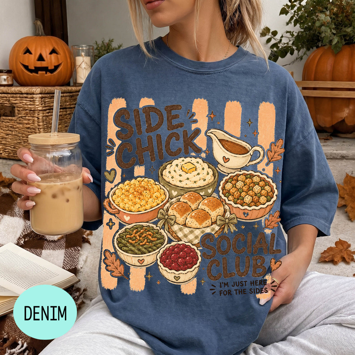 A woman wearing a blue t-shirt with a graphic design of various dishes and a pumpkin, holding a drink and surrounded by autumn decorations.