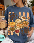 A woman wearing a blue t-shirt with a graphic design of various dishes and a pumpkin, holding a drink and surrounded by autumn decorations.