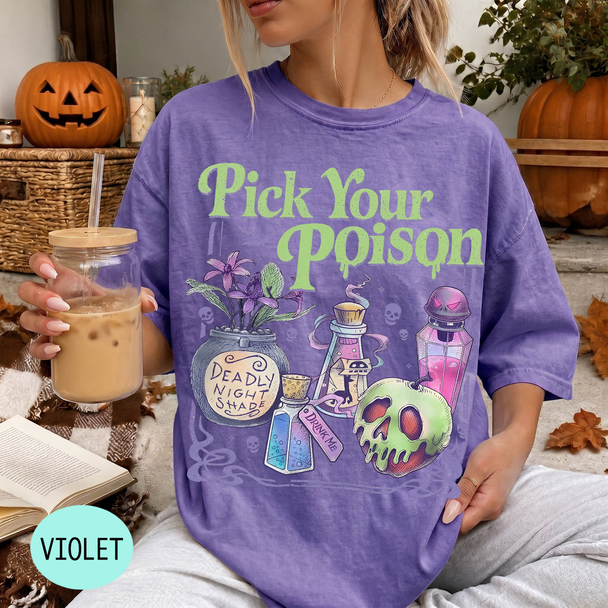 A woman wearing a purple t-shirt with a Halloween-themed design, holding a drink and surrounded by Halloween decorations.