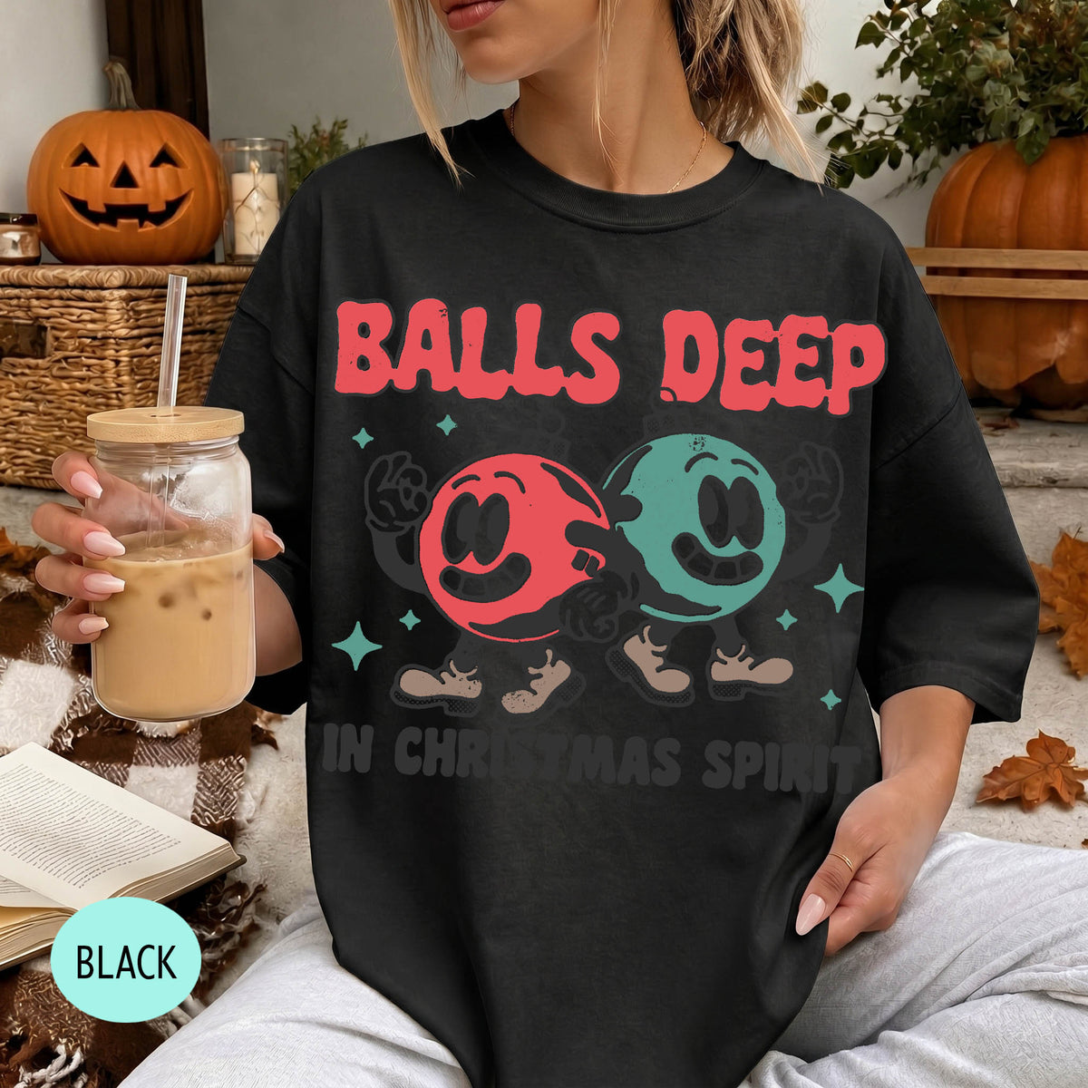 A person wearing a black t-shirt with a graphic design of two cartoon characters and the text "BALLS DEEP" printed on it, holding a drink.
