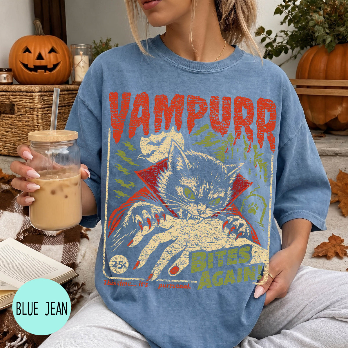 A woman wearing a blue t-shirt with a graphic design of a cat and the text "VAMPURR BITE'S AGAIN" is sitting on a couch with a pumpkin and a book in the background.
