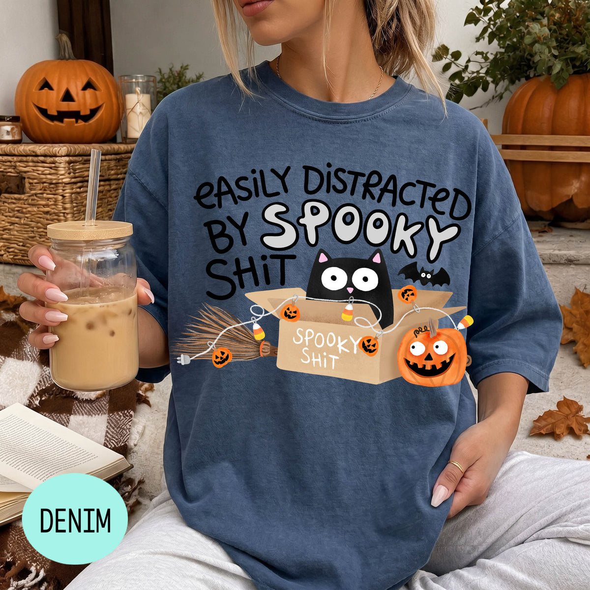 A woman wearing a blue t-shirt with a humorous Halloween-themed graphic, including a cat, pumpkins, and the text "EASILY DISTracted BY SPOOKY SHIT". She is holding a drink in a glass with a straw.