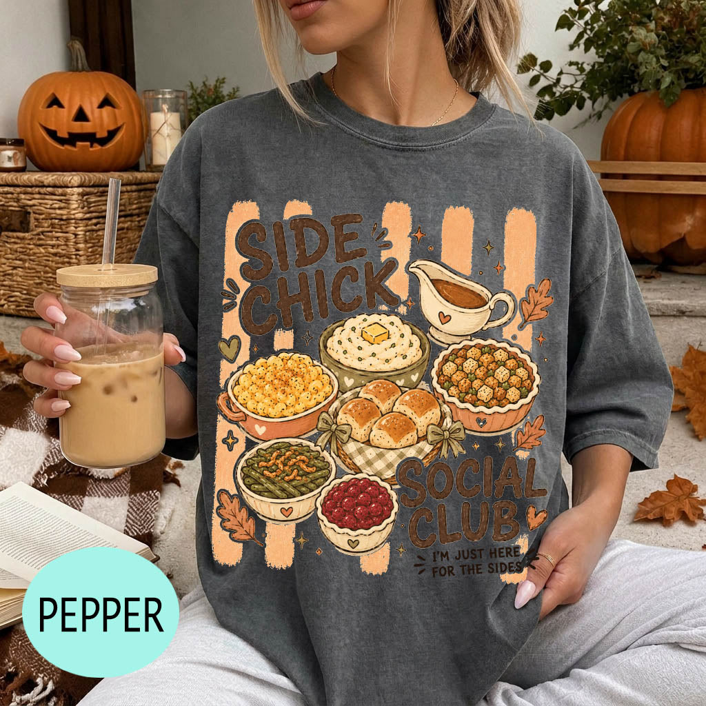 A woman wearing a gray t-shirt with a graphic design of various fall dishes and a pumpkin, holding a drink and surrounded by autumn decorations.